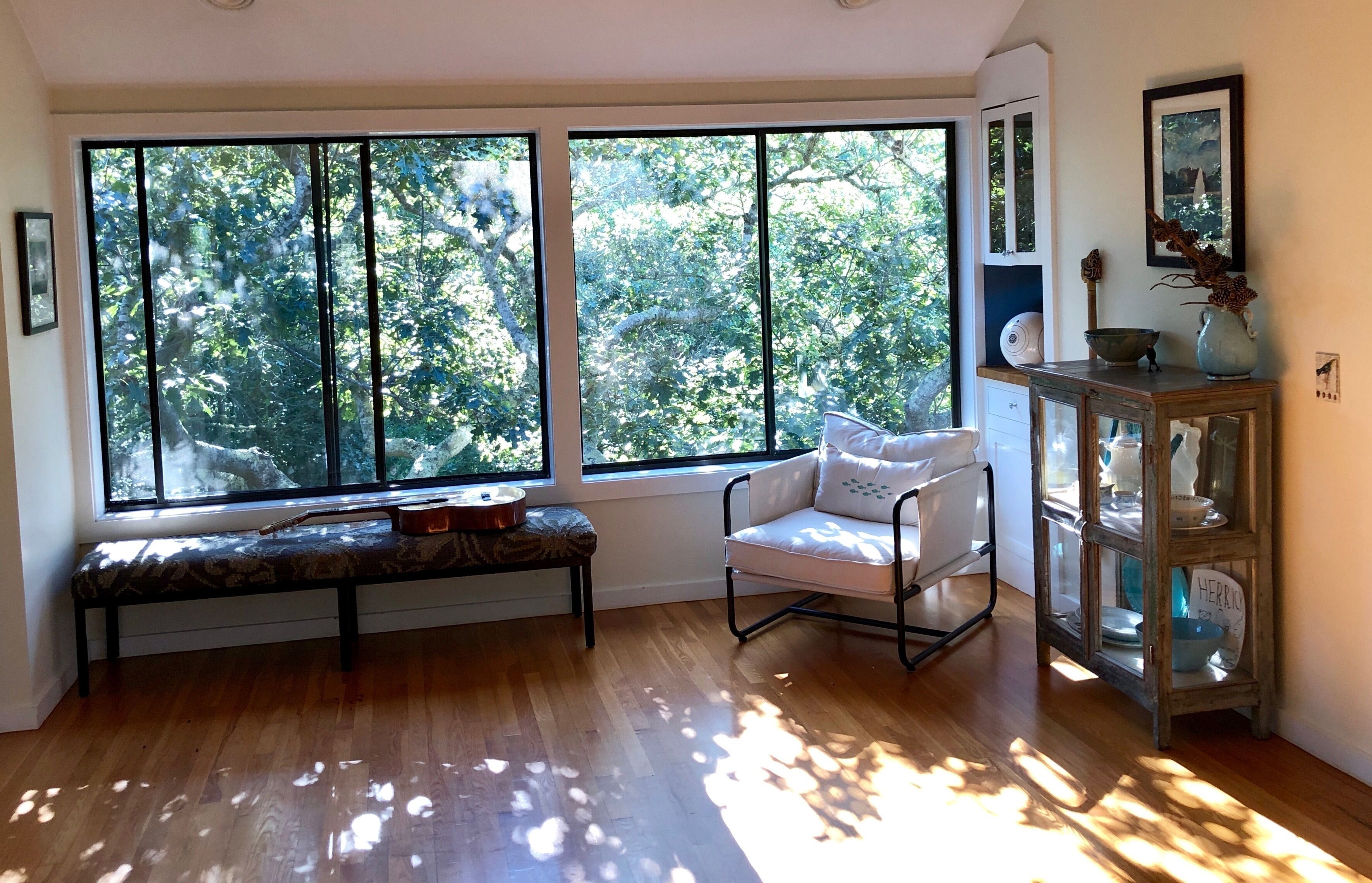 Una habitación con amplios ventanales, un banco, una silla blanca y un aparador de vidrio con objetos decorativos.
