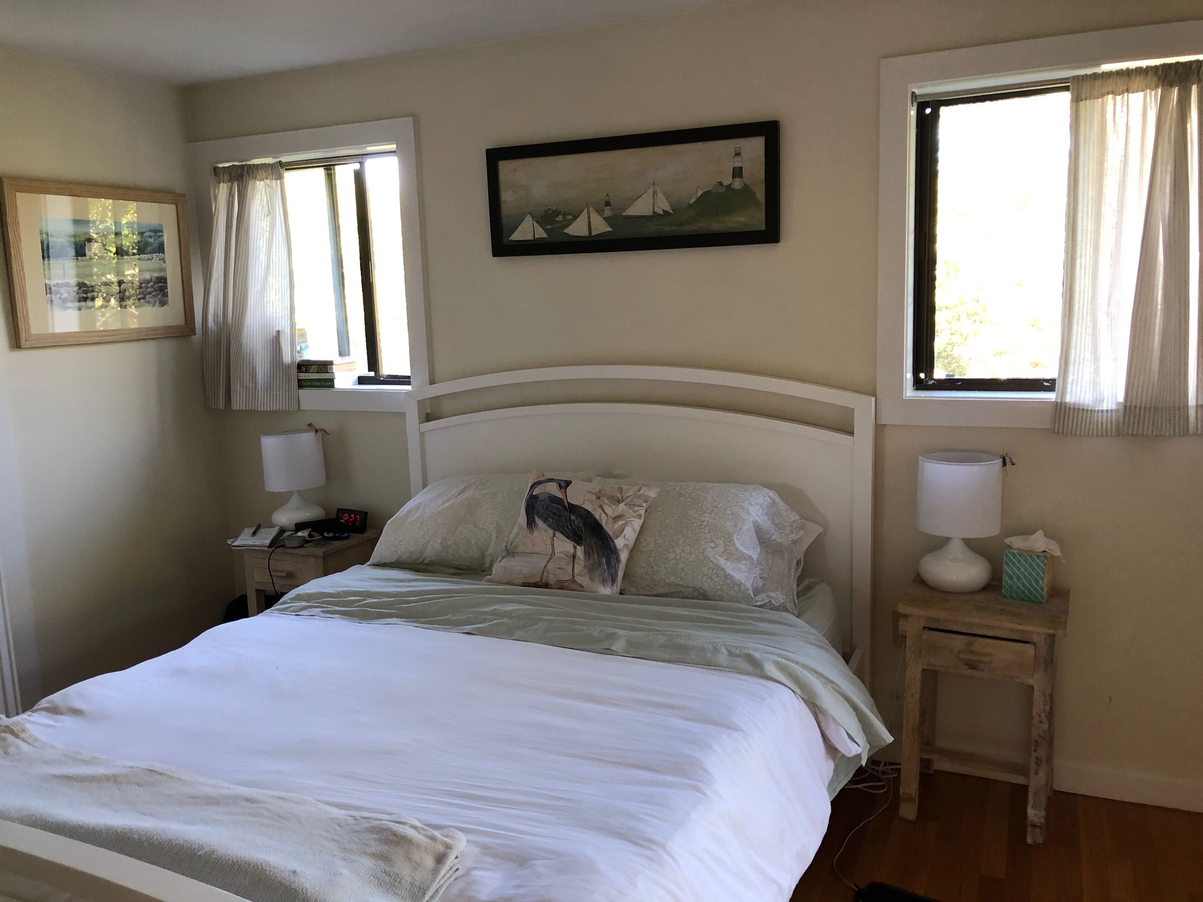 Un lit bien fait, avec un oreiller orné d’un motif d’oiseau, une lampe de chevet, une table de nuit avec un téléphone et une horloge, et un tableau accroché au mur.