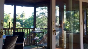 A covered outdoor area with a wooden deck, two chairs, and a small table with glasses and a pitcher. - Magical House in the Trees with Water View - Secluded Area (Aquinnah)