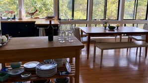 A dining area with a wooden table, wine bottle, and glasses, large windows with a view of trees, and a kitchen area with a sink and decorative items. - Magical House in the Trees with Water View - Secluded Area (Aquinnah)