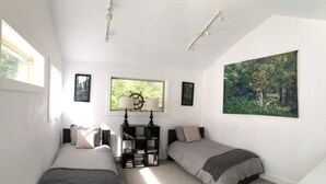 A bedroom with two beds, a bookshelf, a window with a view of greenery, and a painting on the wall. - Chesterman Beach Fine Art Vacation House - Walking distance to North Chesterman (Tofino)