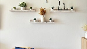 Une pièce avec trois étagères en bois où sont exposées des plantes et des objets décoratifs, une chaise en osier avec un coussin bleu, et une œuvre d’art murale composée de lettres formant le mot « PLAGE ».