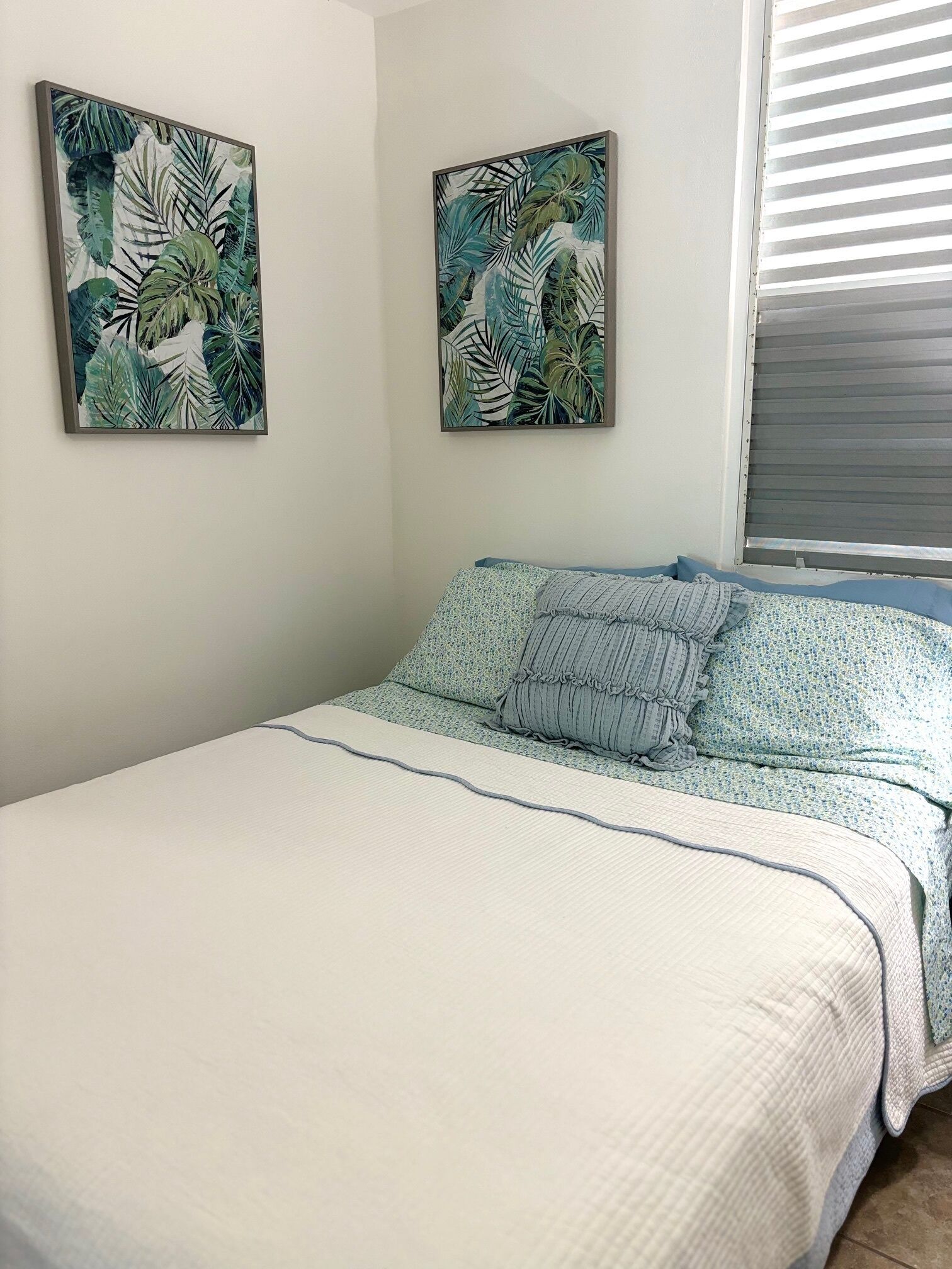 Un dormitorio con una cama, dos cuadros en la pared y una ventana con persianas.