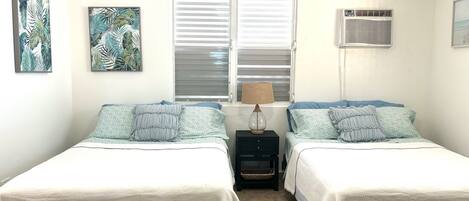 Un dormitorio con dos camas, una ventana con persianas, un aire acondicionado de pared y dos cuadros enmarcados.