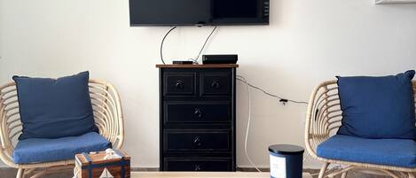 Ein Zimmer mit zwei Korbstühlen mit blauen Kissen, einem Holztisch, einer schwarzen Unterhaltungsecke, einem Wand-Fernseher und einem kleinen Regal mit Pflanzen.