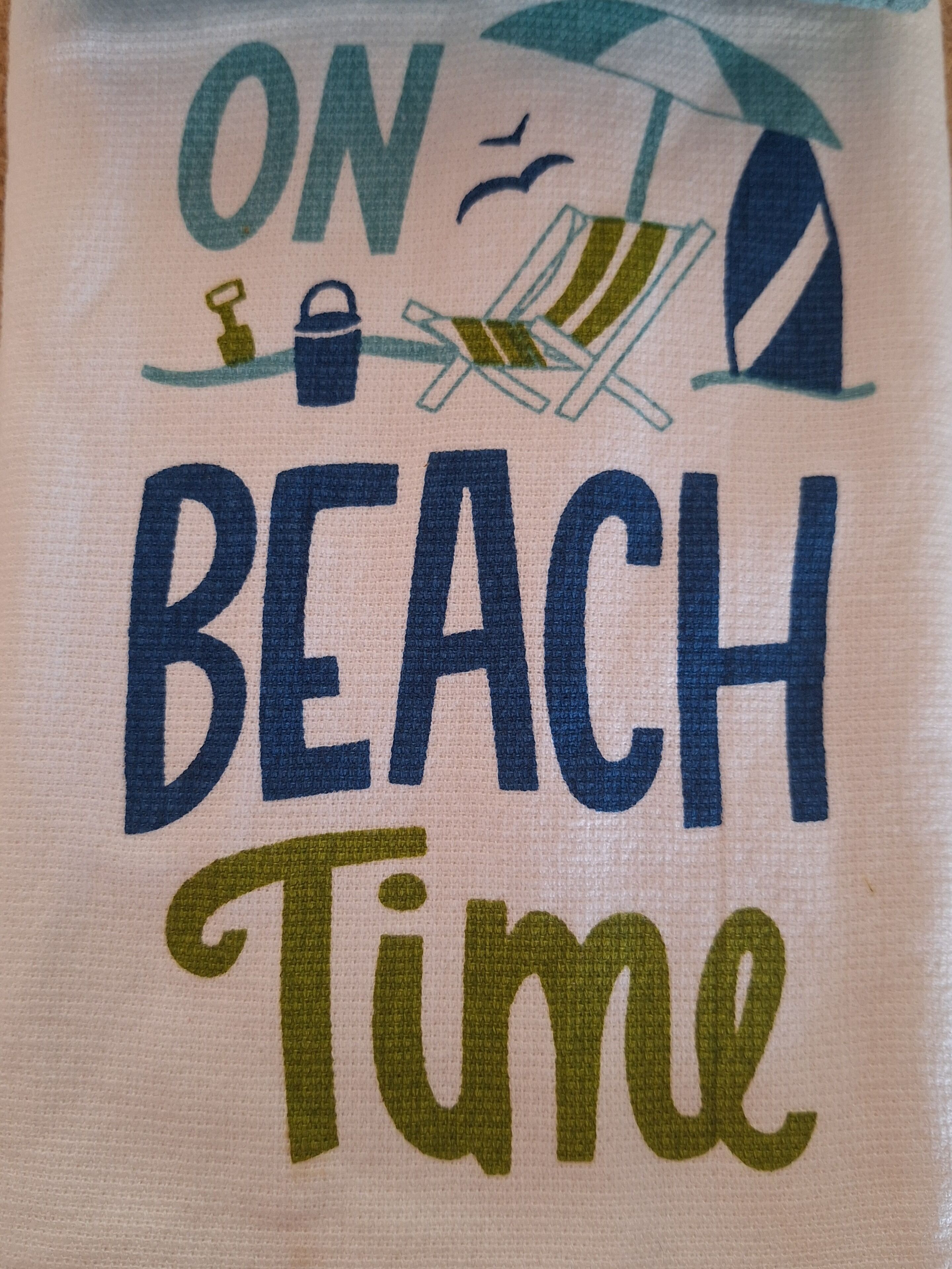 En handduk med texten ”NU ÄR DET STRANDTID” och en illustration av ett strandparasoll och en strandstol.