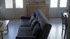 Un salon avec un canapé foncé, une table basse en bois et un coin repas comprenant une table et des chaises. Il y a deux fenêtres avec des rideaux, un lustre et un tableau au mur.