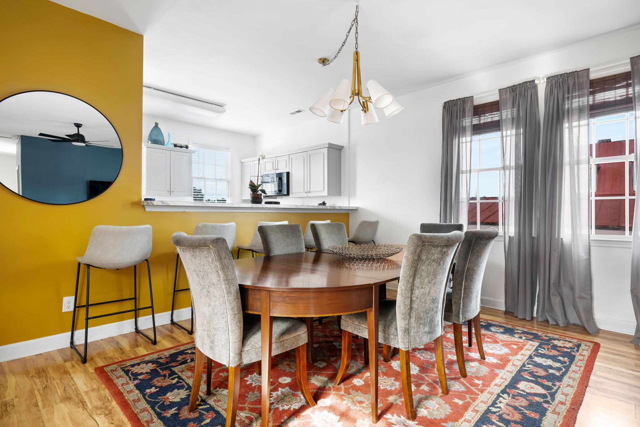 A dining area with a round wooden table, six grey upholstered chairs, a patterned rug, and a modern chandelier.