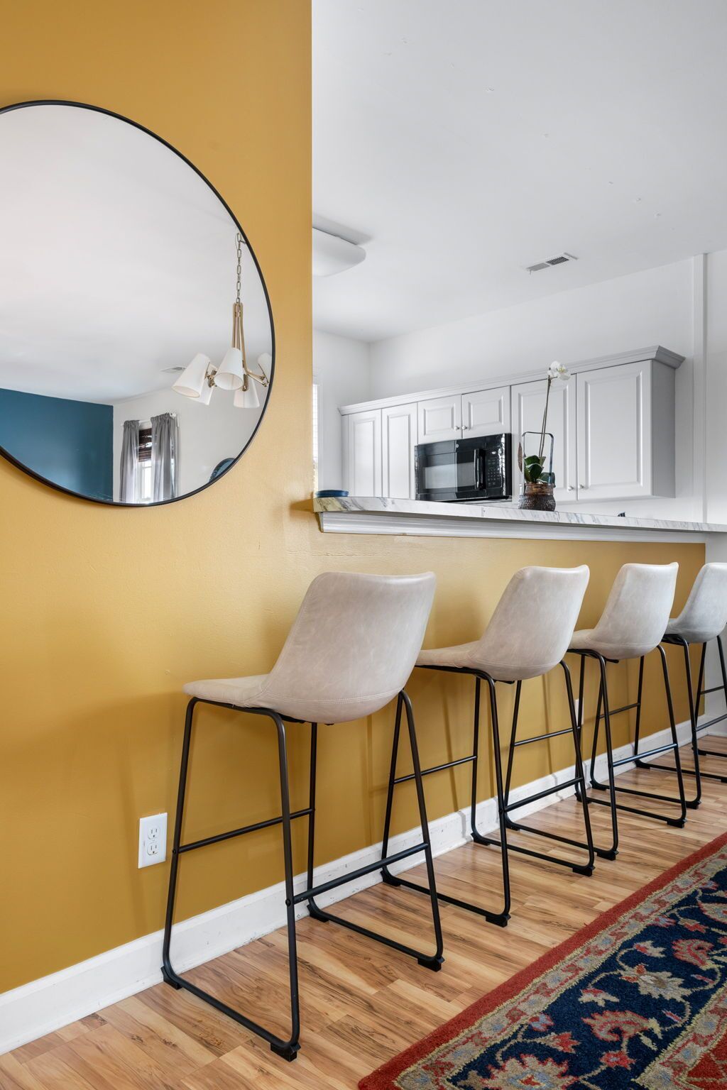 A modern kitchen with white cabinets and a black microwave. There are four bar stools with metal frames and light-colored seats. A round mirror hangs on the wall, reflecting part of the room.