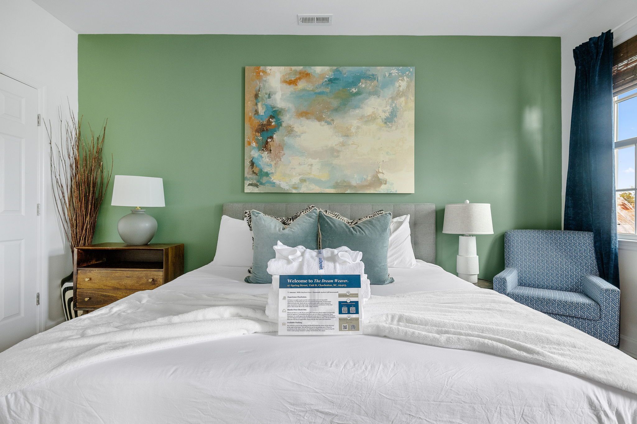 Una cama bien hecha con un cartel de bienvenida, un sillón azul y un cuadro en la pared.