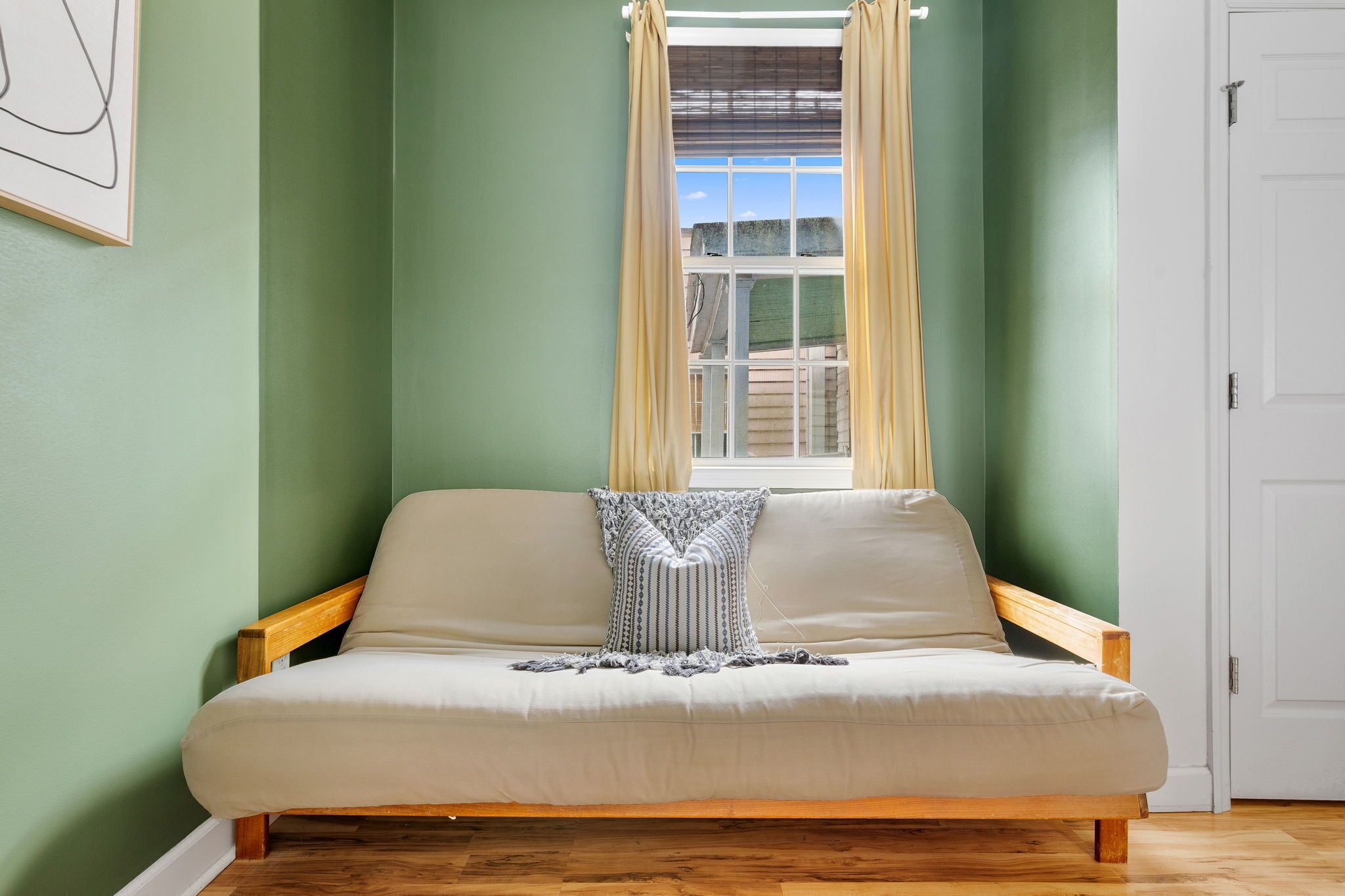 Une pièce avec un canapé-lit placé contre un mur vert, une fenêtre avec des rideaux et une porte blanche.
