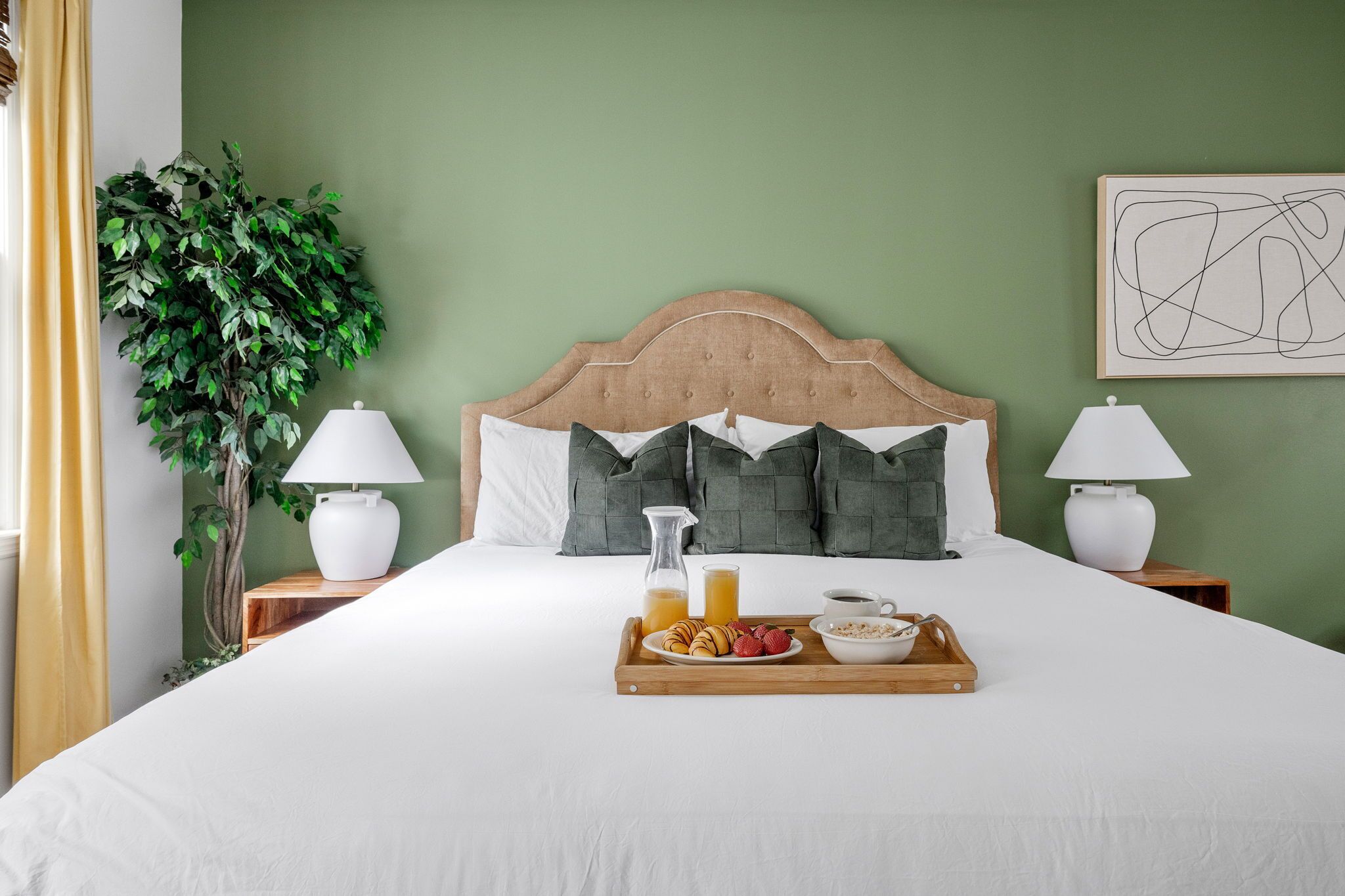 A neatly made bed with a wooden headboard, two bedside lamps, a tray with breakfast items, and a green wall with a framed abstract art piece.