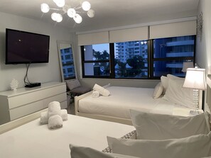 A hotel room with two beds, a TV, a desk, and large windows with a view of buildings.