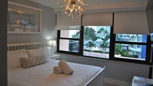 A hotel room with a large bed, a chandelier, a view of palm trees and buildings, and a window with blinds. - Beach Front Resort - 2 Bedroom, 2 bathrooms, Sleeps 6, 2 Pools- at The Alexander (Miami Beach)