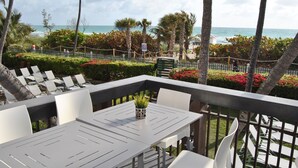 A terrace with white chairs and a table, overlooking a beach with palm trees and a fence. - Beach Front Resort - 2 Bedroom, 2 bathrooms, Sleeps 6, 2 Pools- at The Alexander (Miami Beach)