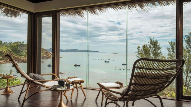 Una habitación con ventanas amplias que dan a una masa de agua, muebles de mimbre y una pequeña mesa con un libro y una taza.