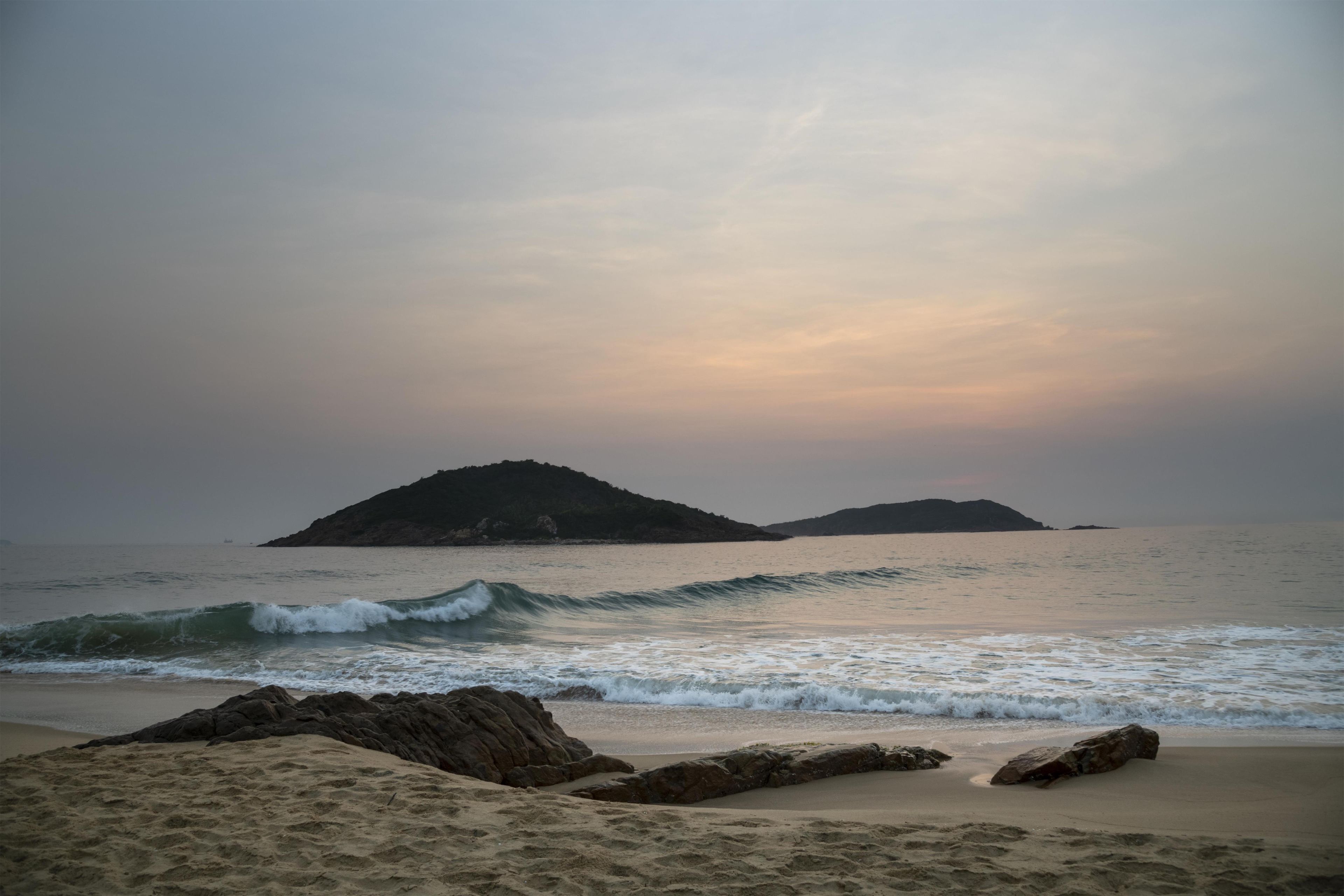 A beach with waves, rocky outcrops, and distant islands under a cloudy sky.