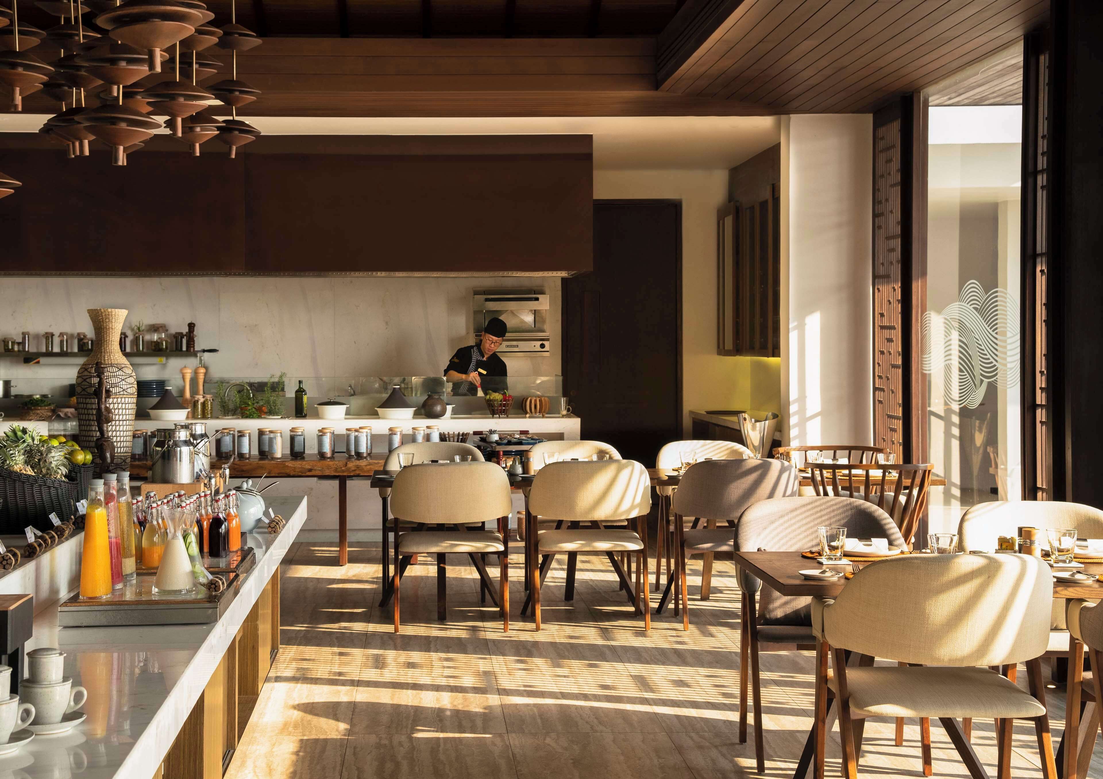 A modern kitchen with a chef preparing food, a bar area with various drinks, and a dining area with wooden chairs and tables.