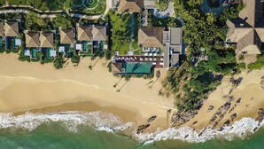 Vista aérea de un complejo turístico con varias construcciones tipo bungalow, un área de piscina central y una playa con olas.
