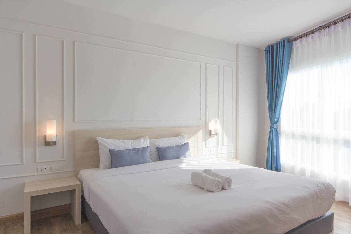A hotel room with a large bed, white walls, and a window with blue curtains.