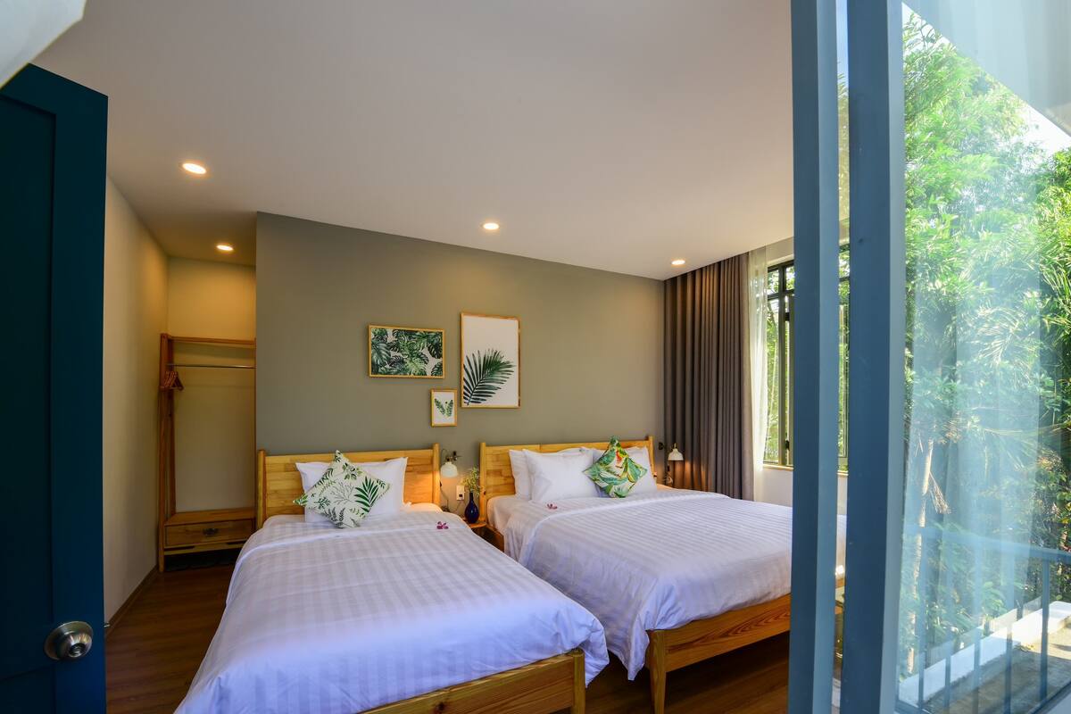 A hotel room with two beds, a wooden wardrobe, a small table with a lamp, and a window with a view of greenery.
