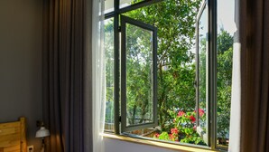 Habitación de hotel con cama, mesita de noche, lámpara y ventana con vistas a follaje y flores.