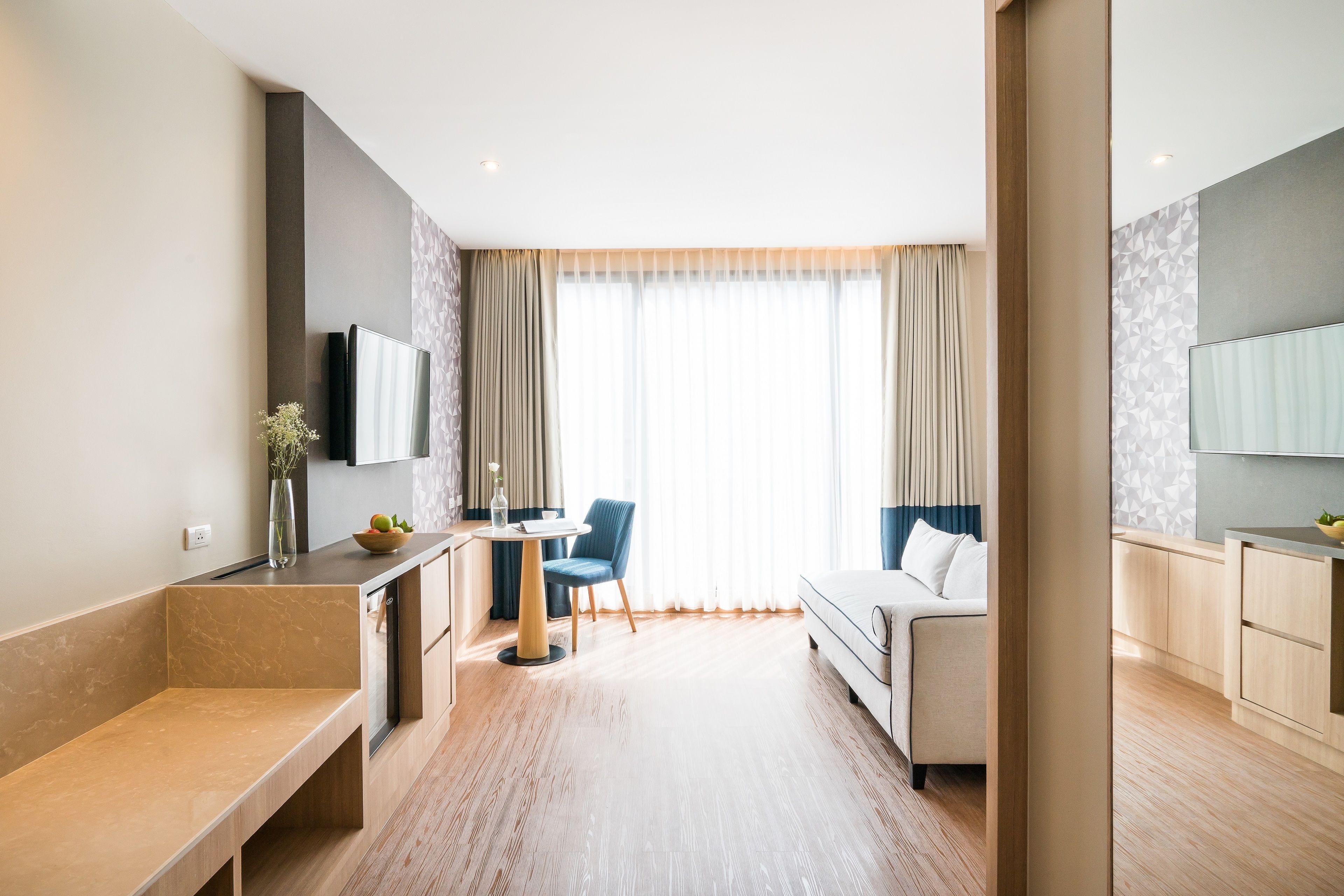 A modern hotel room with a sofa, a small table, a chair, a TV mounted on the wall, and a large window with curtains.