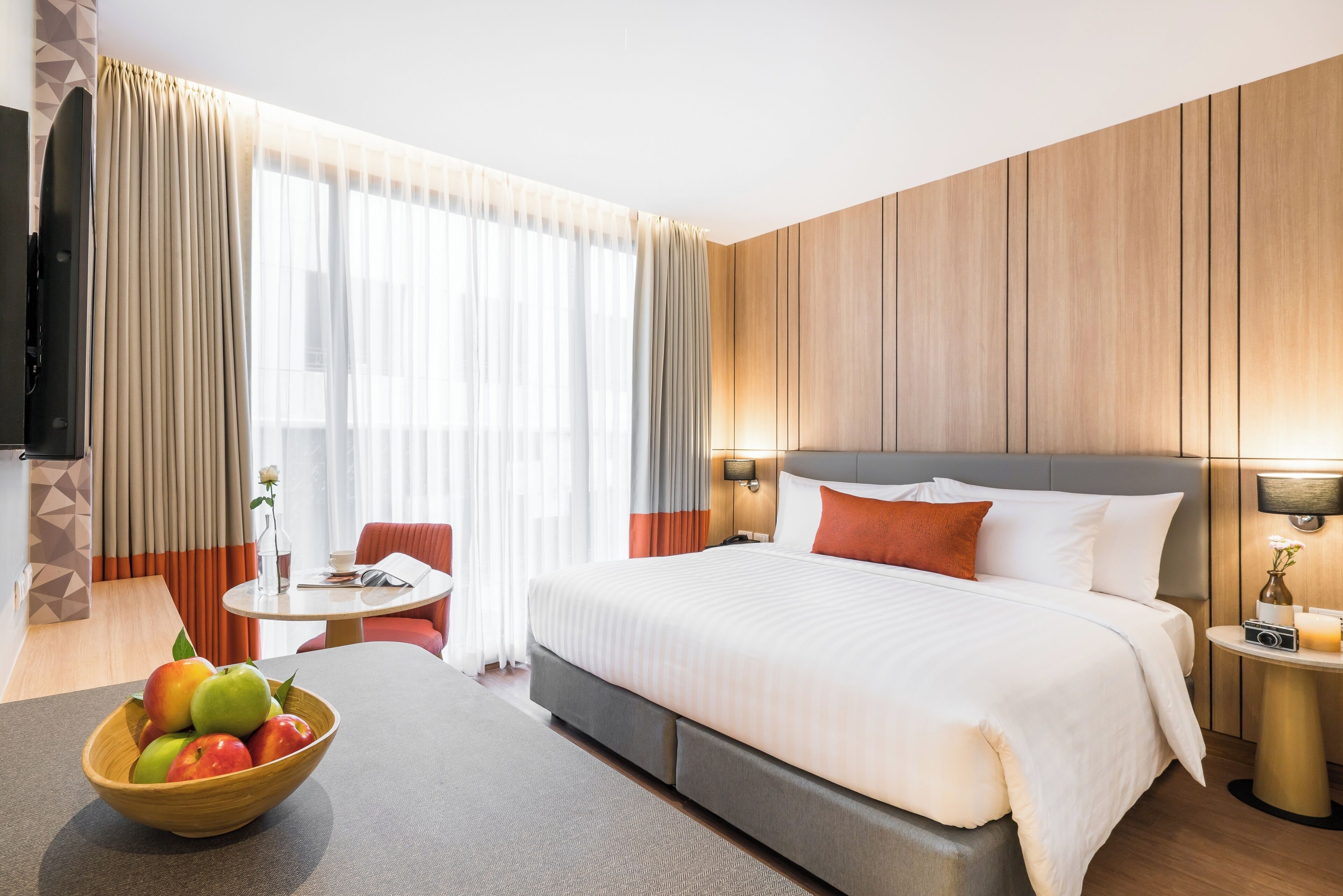 A hotel room with a bed, a dining table, a bowl of fruit, and a TV.