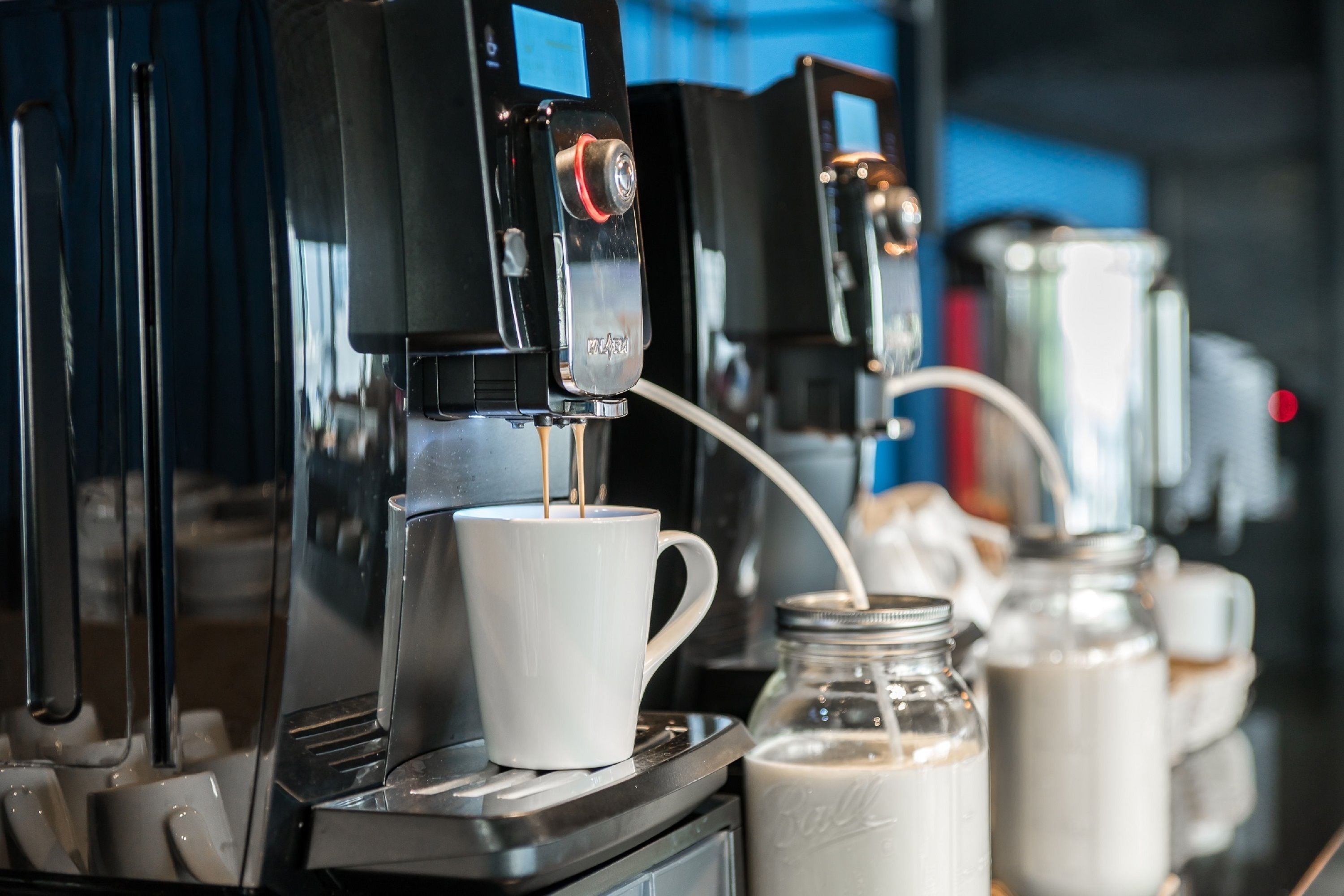 Une machine à café verse du café dans une tasse, avec des bouteilles de lait et d’autres accessoires pour préparer le café en arrière-plan.