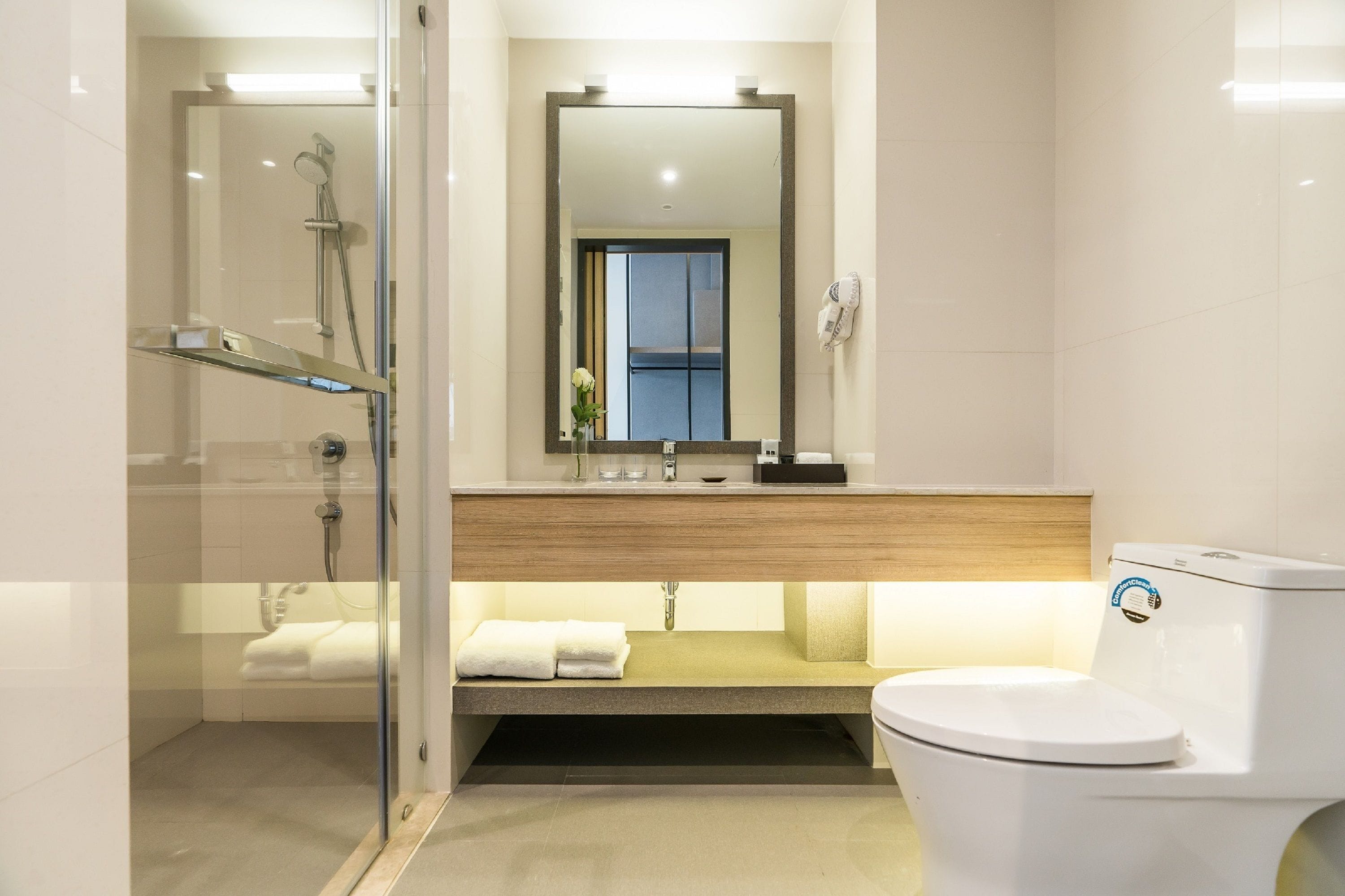 A modern bathroom with a glass-enclosed shower, a wooden vanity, a white toilet, and a large mirror above the sink.