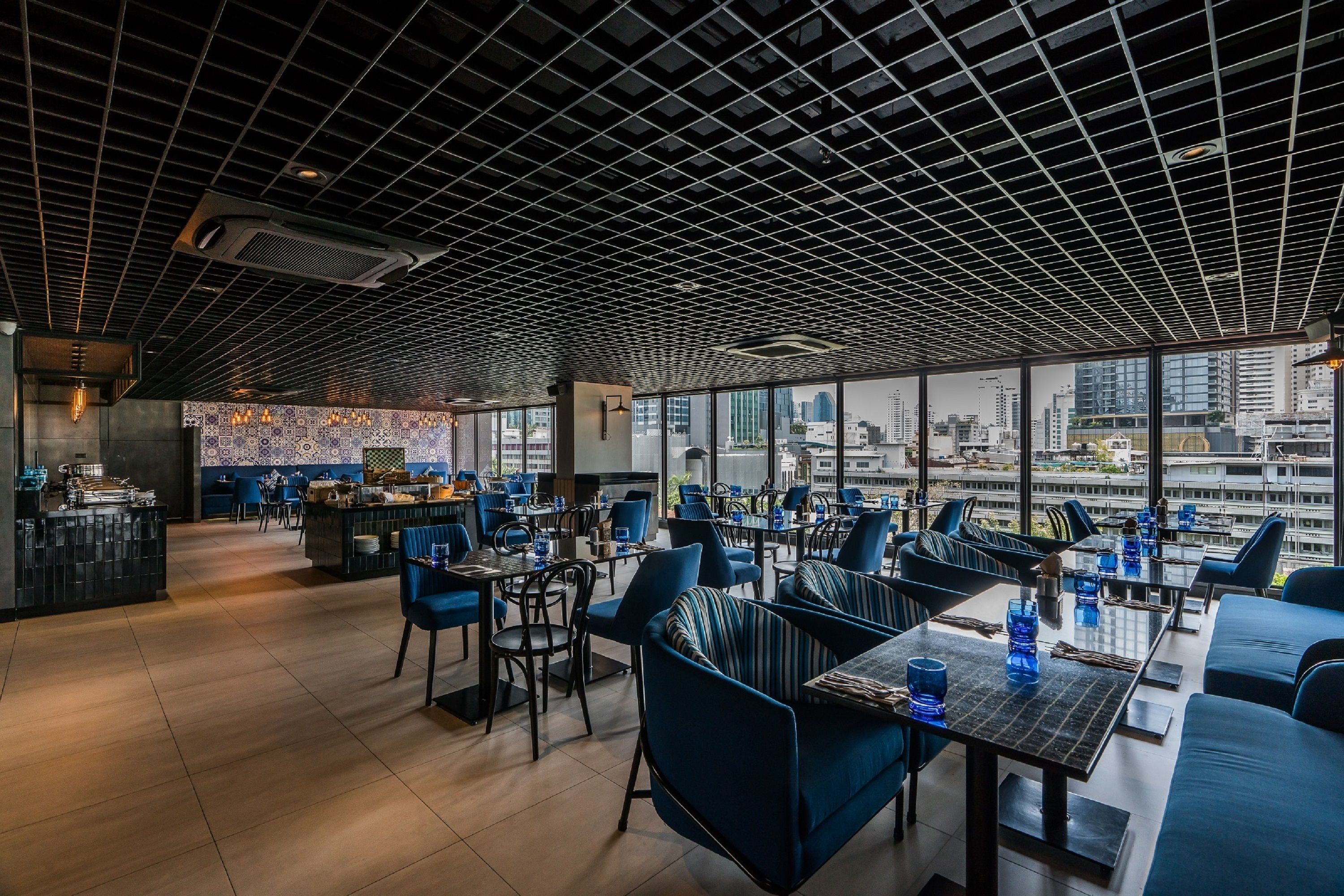 A modern restaurant with a grid-patterned ceiling, large windows offering city views, and blue upholstered seating.
