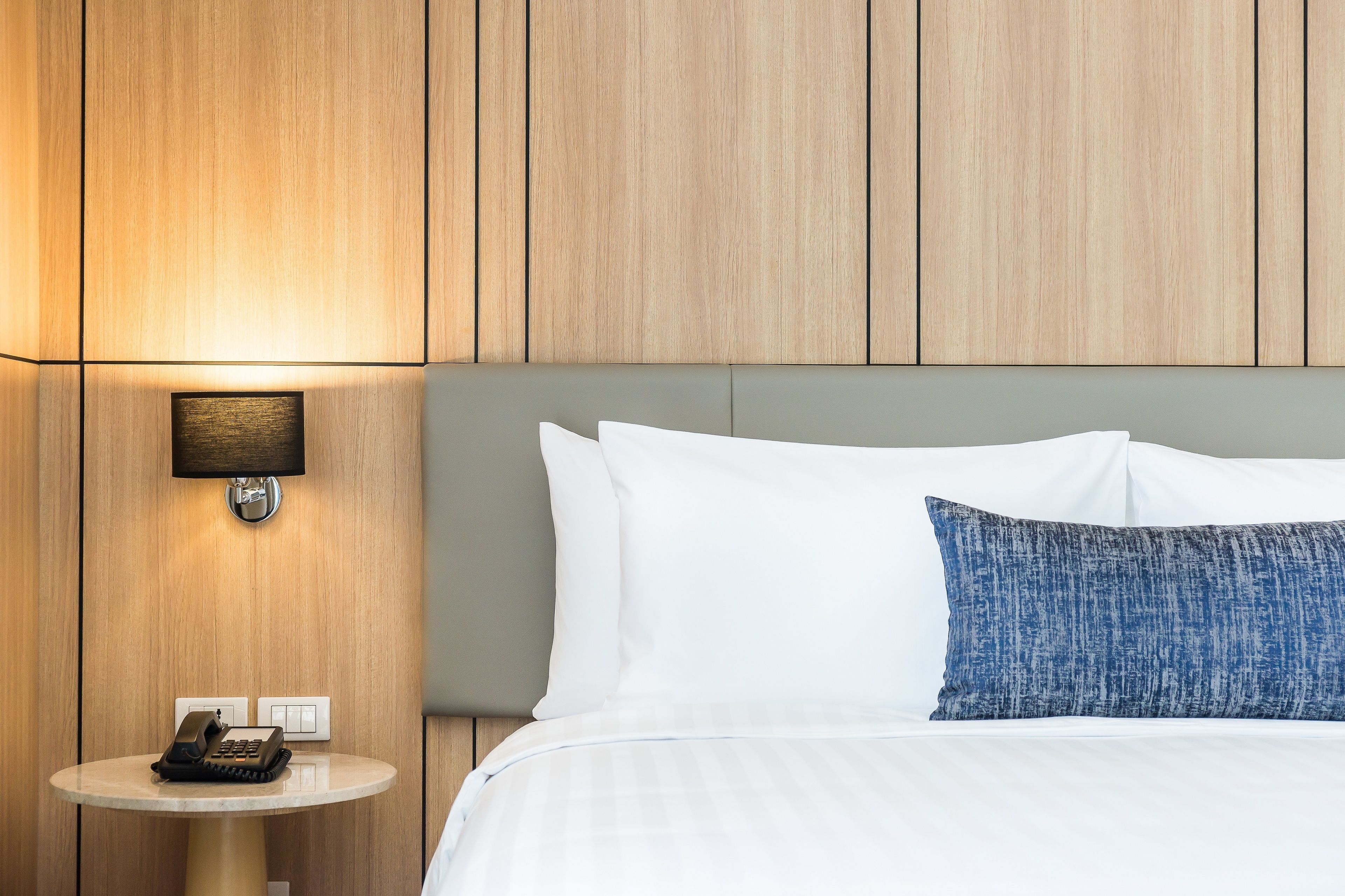 Ein Hotelzimmer mit einer Wand aus Holzpaneelen, einer Nachttischlampe, einem runden Tisch mit Telefon und einem Bett mit blau-weißen Kissen.