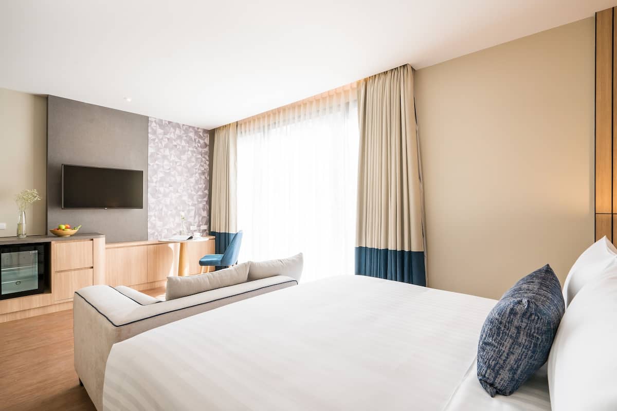 A modern hotel room with a large bed, a flat-screen TV mounted on the wall, a desk with a chair, and a window with curtains.