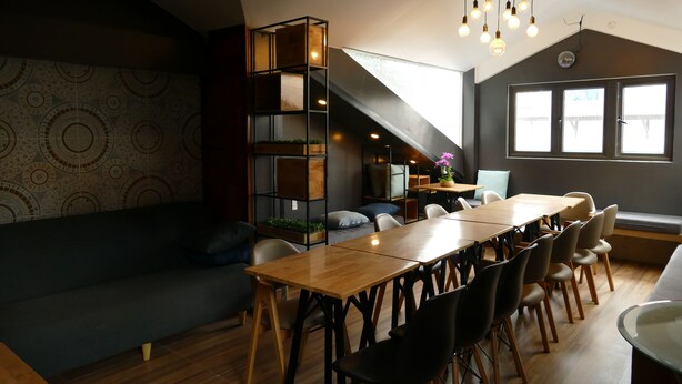 A modern dining area with a wooden table, black chairs, a dark sofa, and a unique shelving unit with plants. - Dalat Sky Hostel (Xuan Huong - Da Lat)
