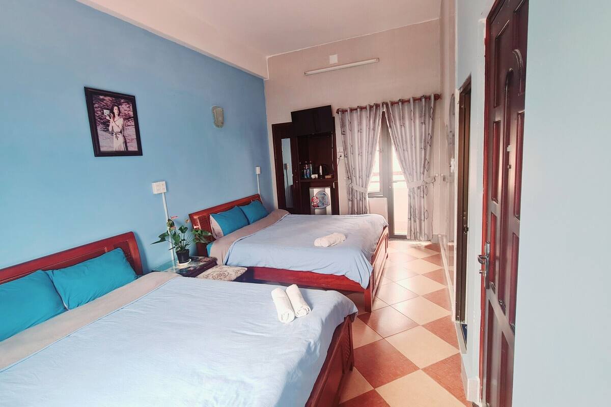 Habitación de hotel con dos camas, una mesita con una planta, un cuadro en la pared y una puerta que da a otra habitación.