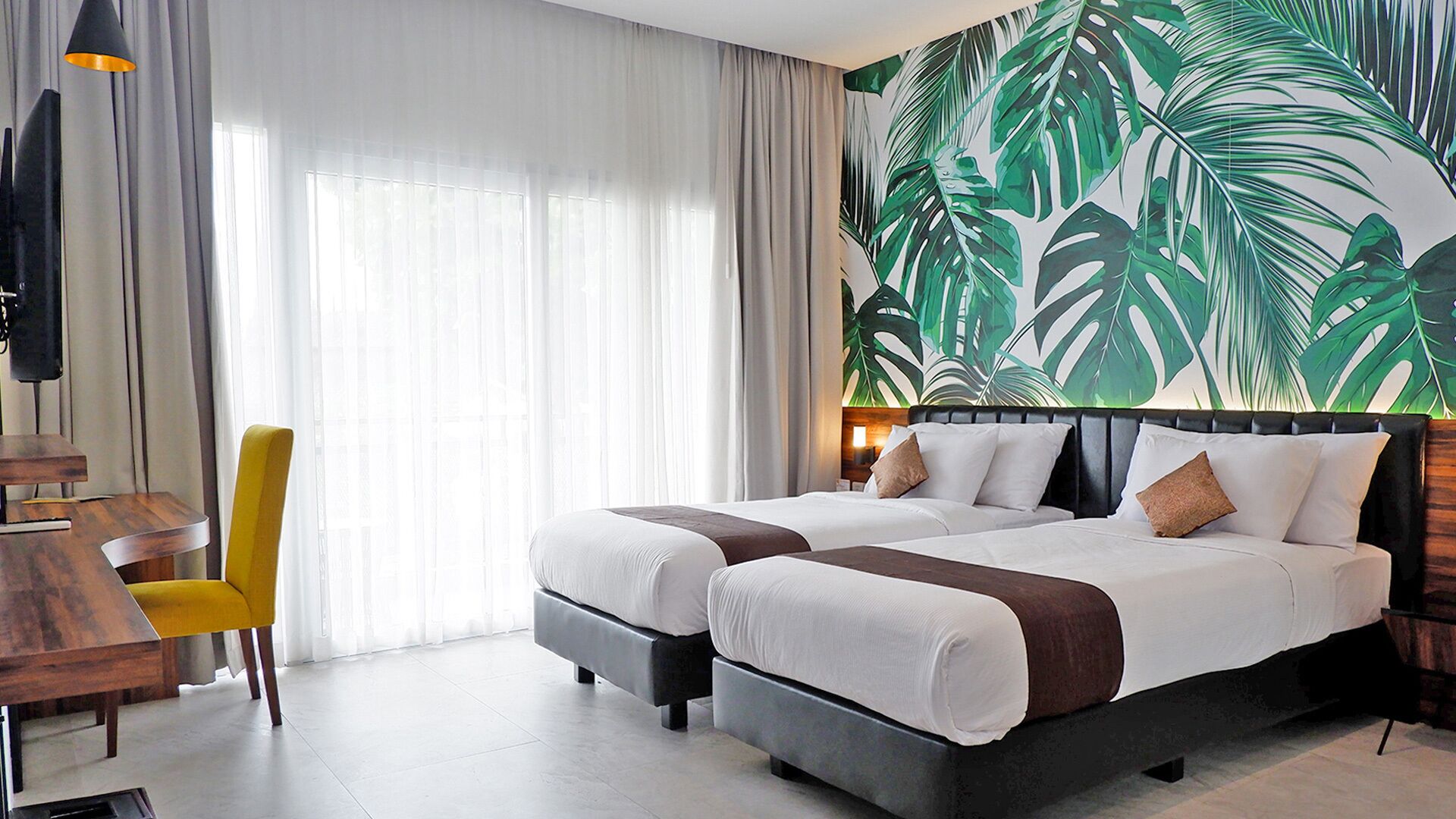 Camera d'albergo con due letti, una scrivania, una sedia e un grande murale con foglie tropicali.