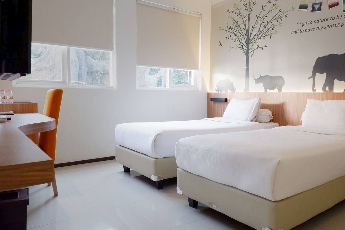 Ett hotellrum med två sängar, ett skrivbord och en stol. På en av väggarna finns en väggmålning med ett träd, en ko och en buffel.