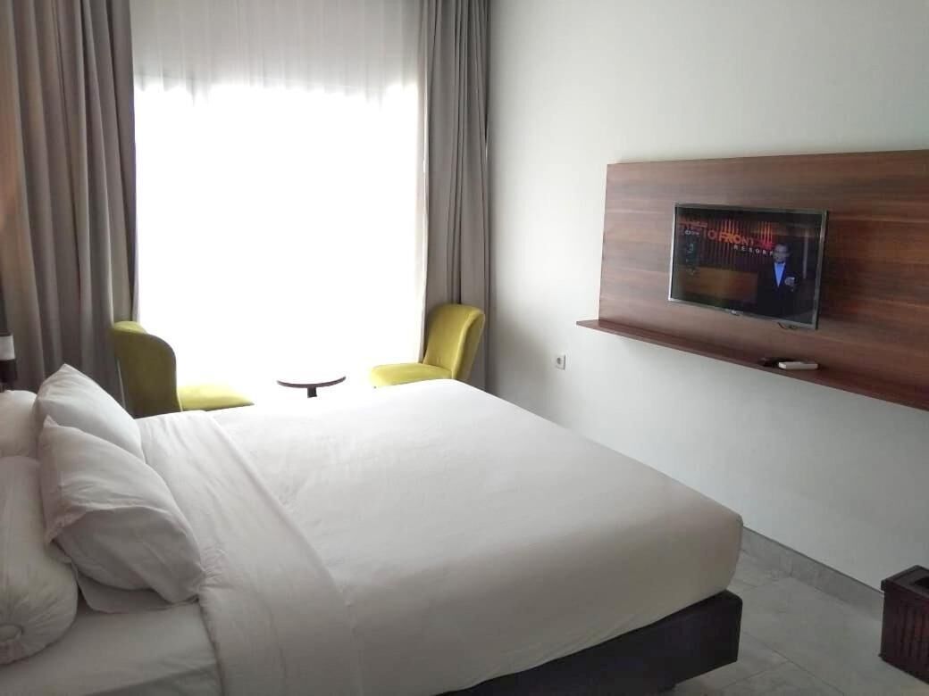 A hotel room with a large bed, a TV mounted on the wall, a small table, and two yellow chairs.