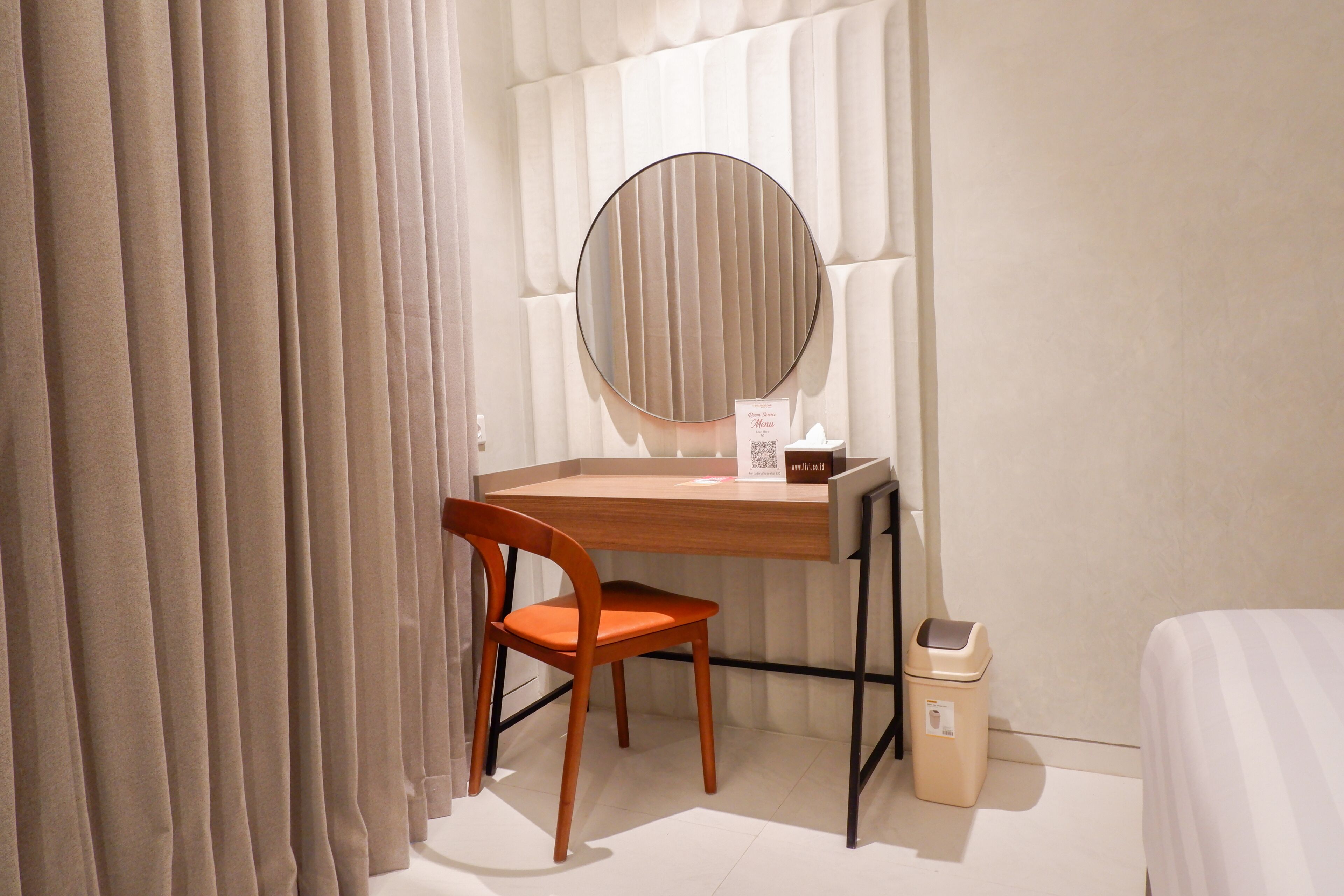 Una habitación de hotel con un escritorio de madera, un espejo redondo, una silla, un pequeño cubo de basura y una cama.