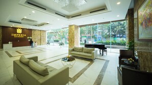 A spacious lobby with a reception desk, sofas, a piano, and a large window offering a view of the outdoors. - Vistay (Hanoi)