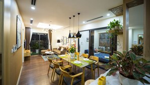 A modern dining area with a wooden table, chairs, and a vase of flowers. There is a sofa and a window with curtains in the background. - Vistay (Hanoi)