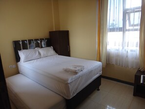 A neatly made bed with white linens and a folded towel, a dark wooden headboard, and a window with sheer curtains. - Front One Inn Malang (Malang)