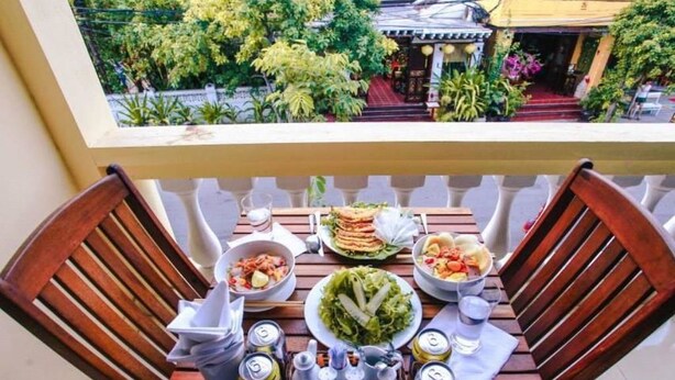A wooden table with food and drinks on a balcony with a view of a traditional building and greenery. - Harmony Homestay (Da Nang)