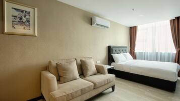 Quarto de hotel com sofá bege, cama com roupa branca, ar condicionado e quadro pendurado na parede.