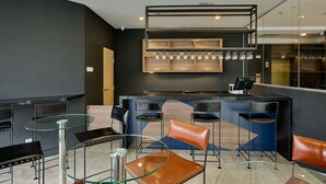 Zona de bar moderna con una mesa de vidrio, sillas de cuero y una estantería de madera con vasos.