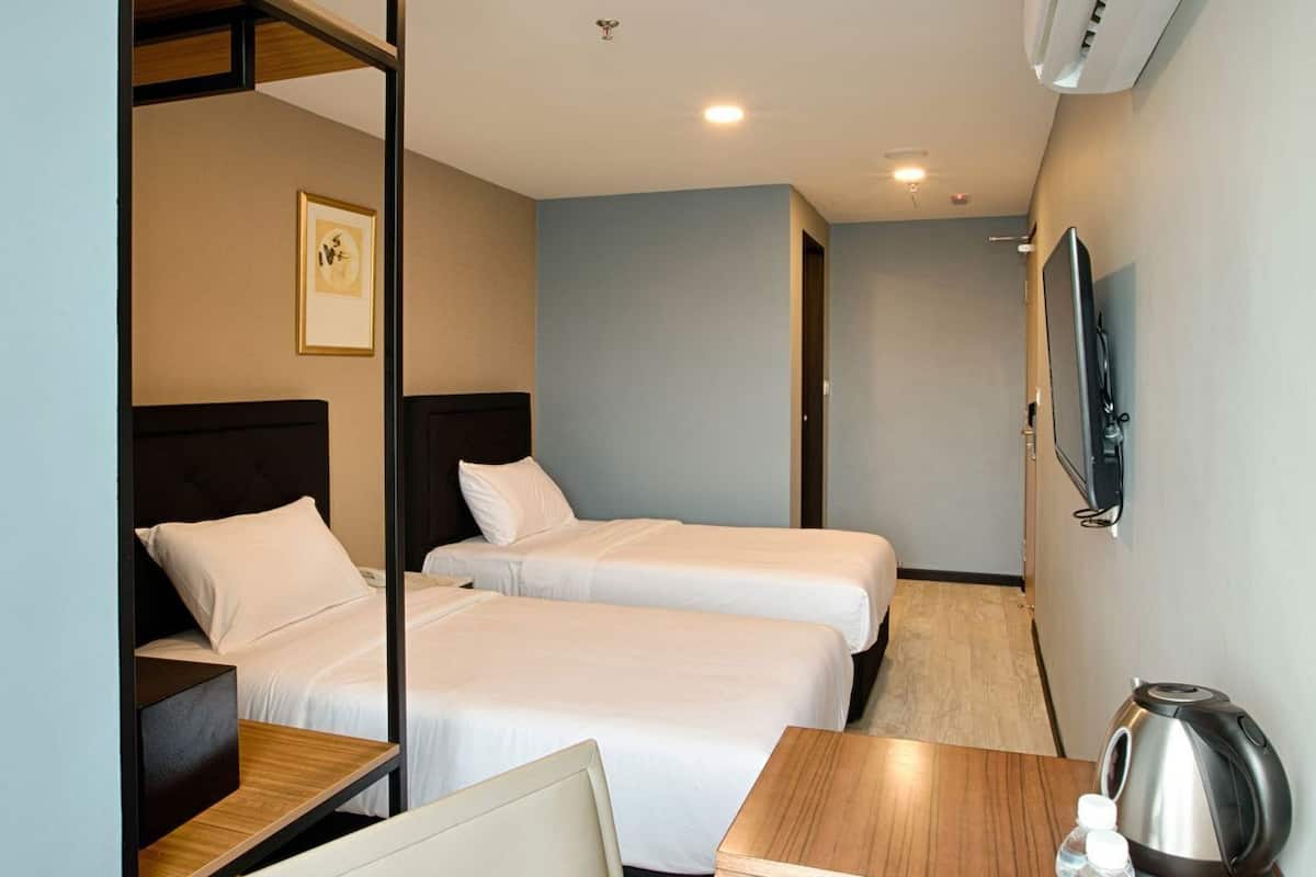 Une chambre d’hôtel avec deux lits, un bureau en bois, une chaise, une bouilloire et une bouteille d’eau.