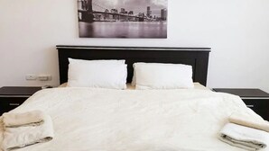 Un lit bien fait, avec des draps blancs et deux oreillers, une tête de lit noire et une photographie représentant un paysage urbain accrochée au mur.