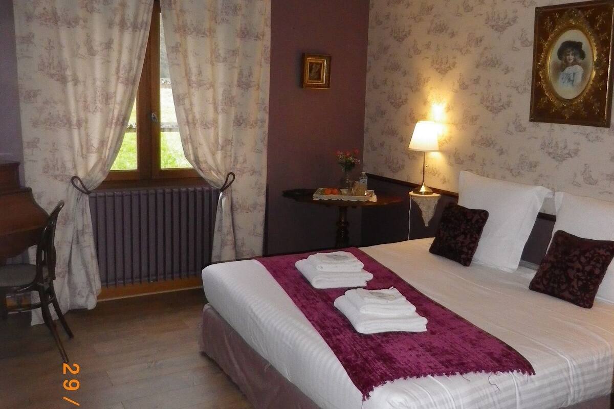 Une chambre d’hôtel avec un lit, un bureau, une chaise, une fenêtre avec des rideaux, une lampe et un tableau encadré au mur.