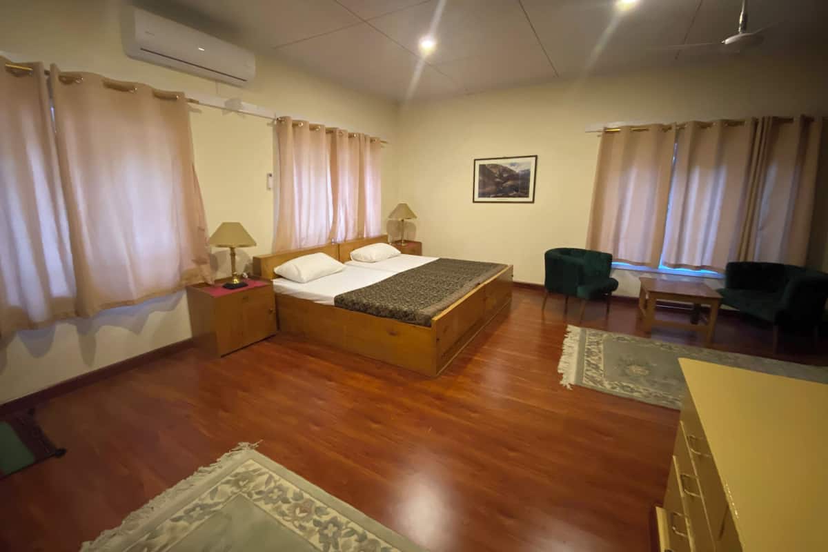 Un dormitorio con una cama de madera, dos mesitas de noche con lámparas, un sillón verde y una ventana con cortinas.