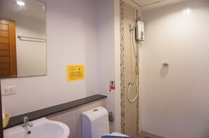 Baño con lavamanos, inodoro y ducha. Hay un letrero amarillo en la pared y un espejo sobre el lavamanos.